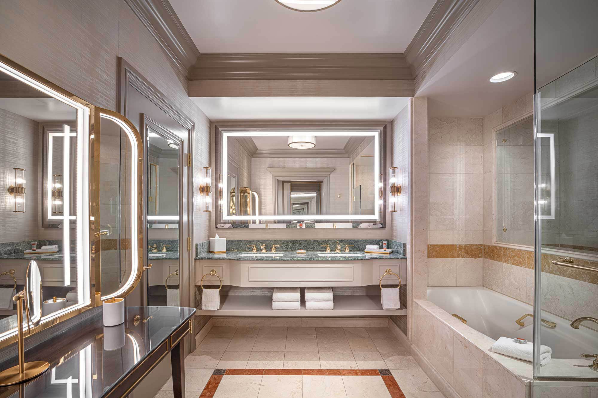 Luxurious bathroom with large mirrors, a spacious vanity, beige tiled walls, a glass shower, and a bathtub.