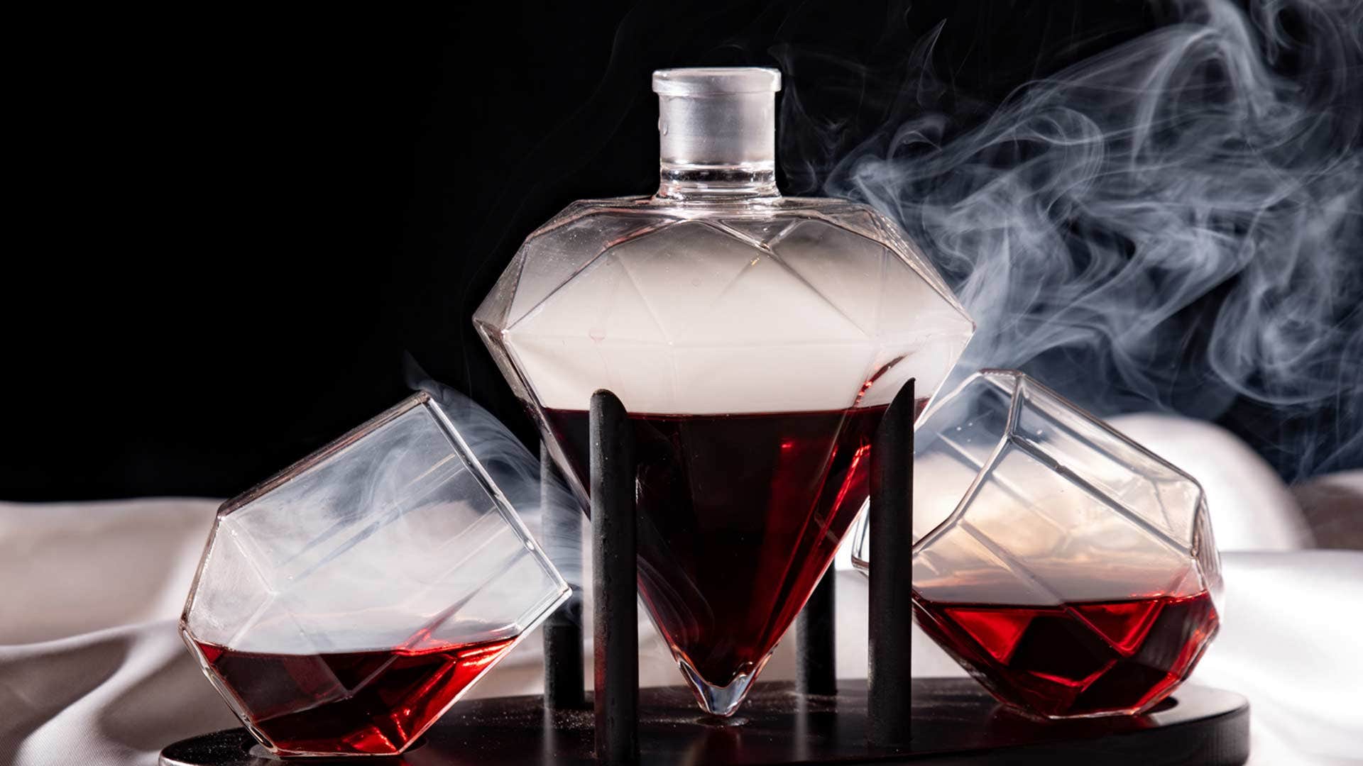 A diamond-shaped decanter with red liquid and smoky top, flanked by two matching tilted glasses filled halfway.