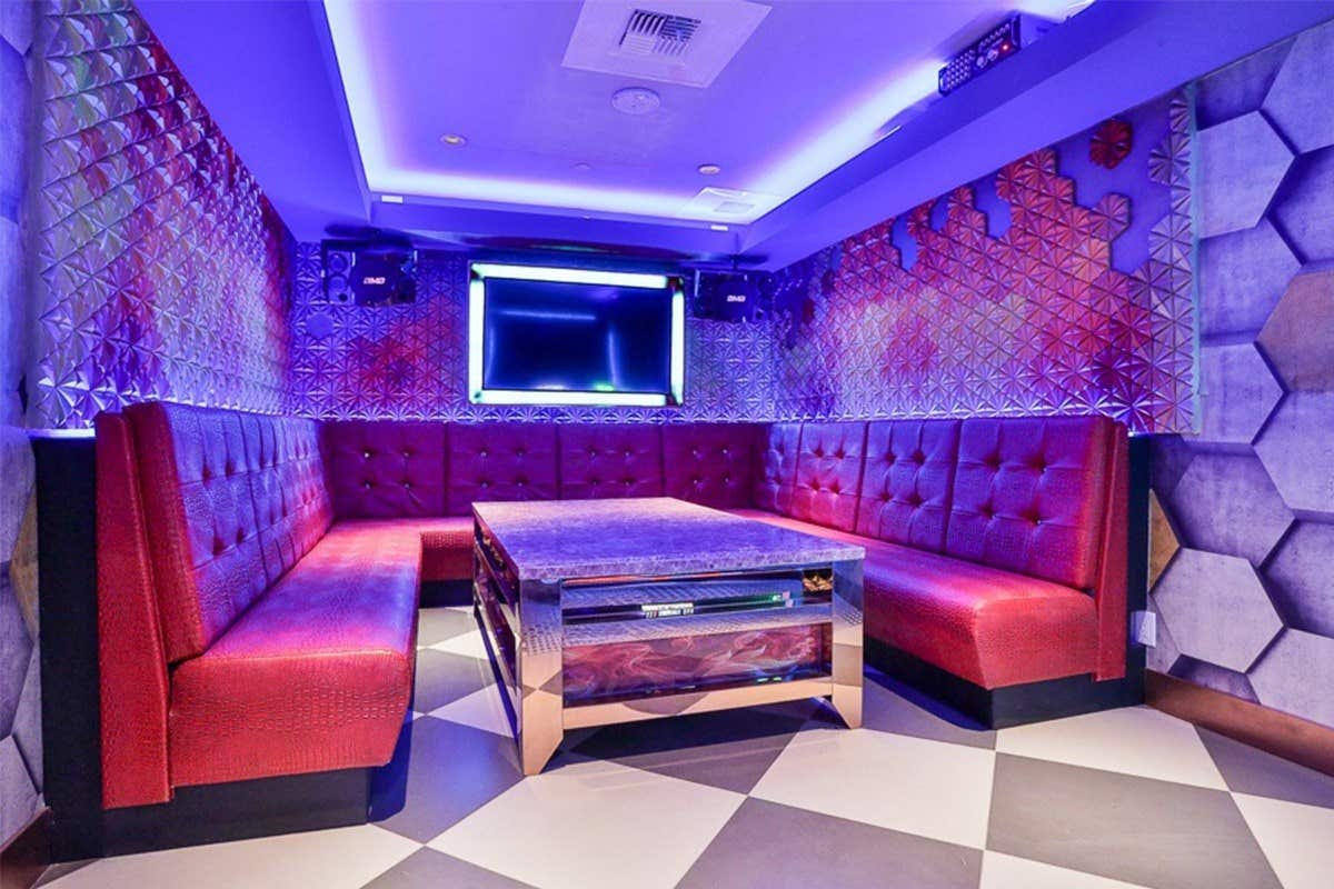 A modern karaoke room featuring purple lighting, plush red seating, a central table, and a large screen on the wall.