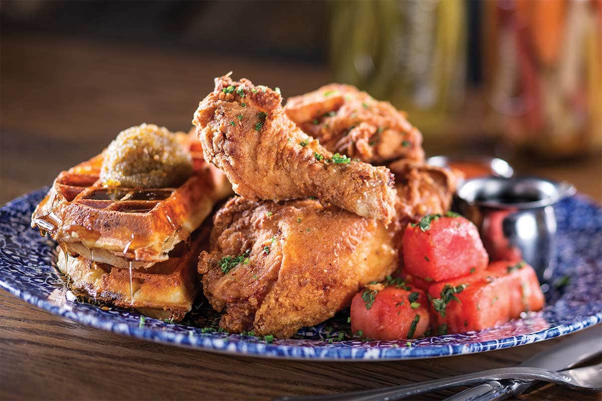 A plate with fried chicken, waffles, watermelon pieces, and a side of sauce on a patterned dish.