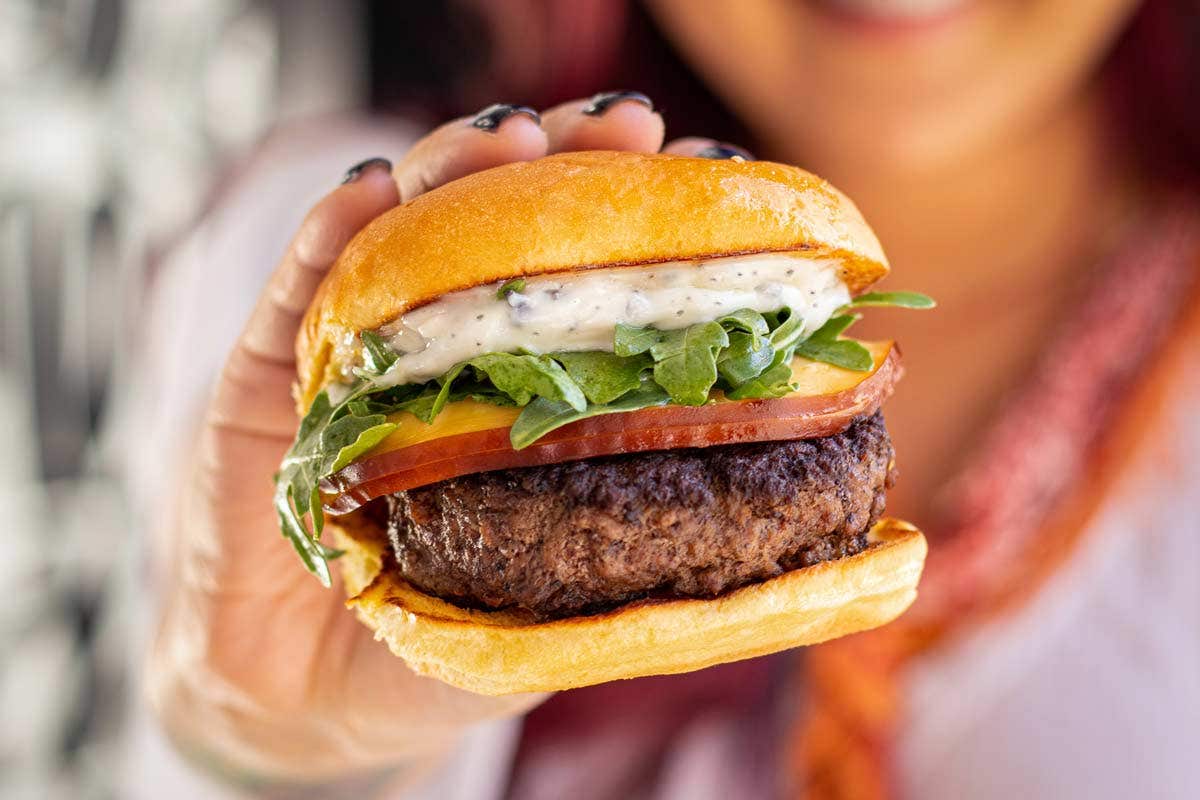 Person holding a burger with lettuce, tomato, arugula, sauce, and a beef patty in a bun.