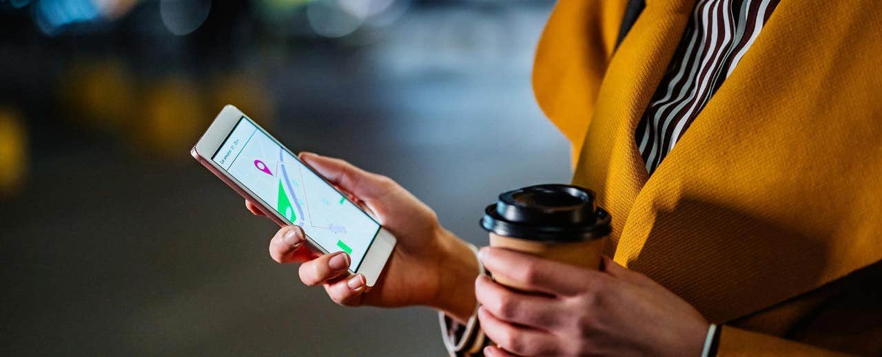 Person using a smartphone with a GPS app while holding a coffee cup, wearing a yellow coat.