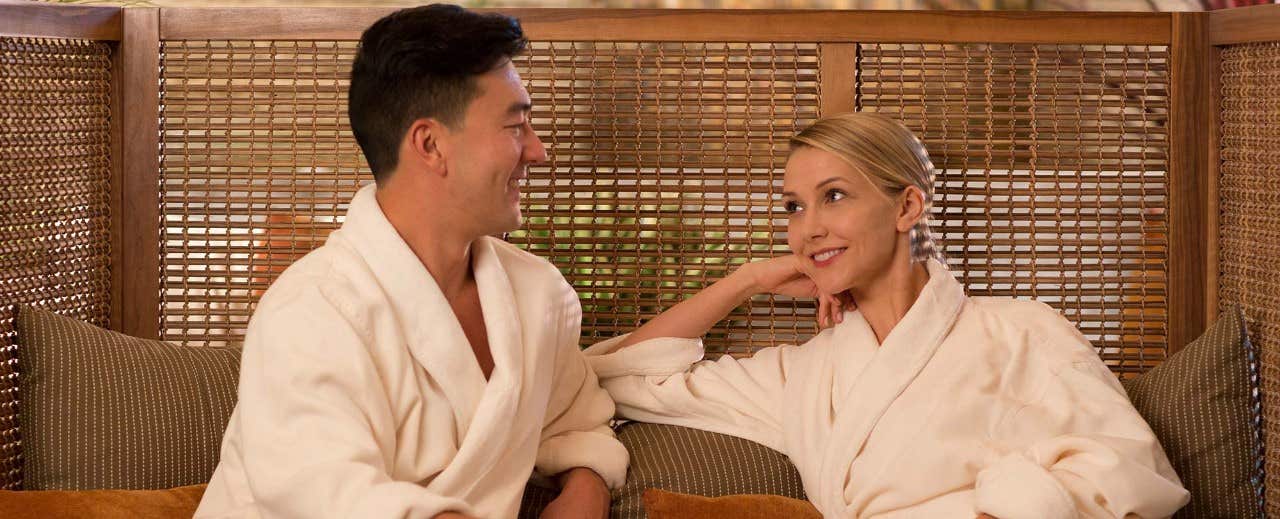 A smiling couple wearing white robes sitting on a couch, talking and relaxing with a wicker background.