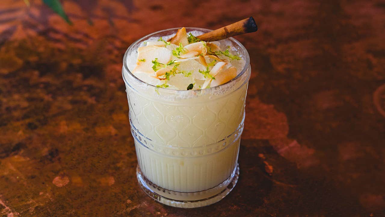 A creamy cocktail in a textured glass, garnished with lime zest, toasted coconut flakes, and a cinnamon stick.