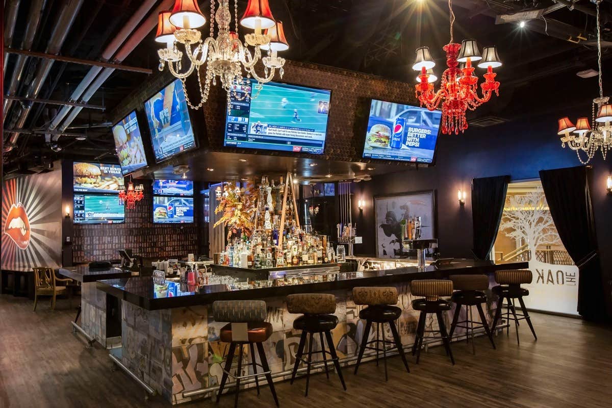 Modern sports bar with multiple TV screens, a central bar surrounded by stools, and eclectic chandeliers overhead.