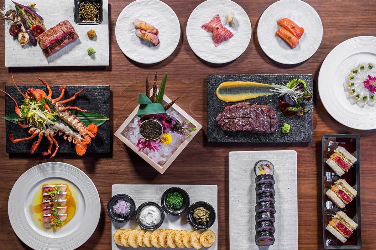 An overhead shot of various plates of gourmet Japanese cuisine on a wooden table, including sushi, steak, and seafood.