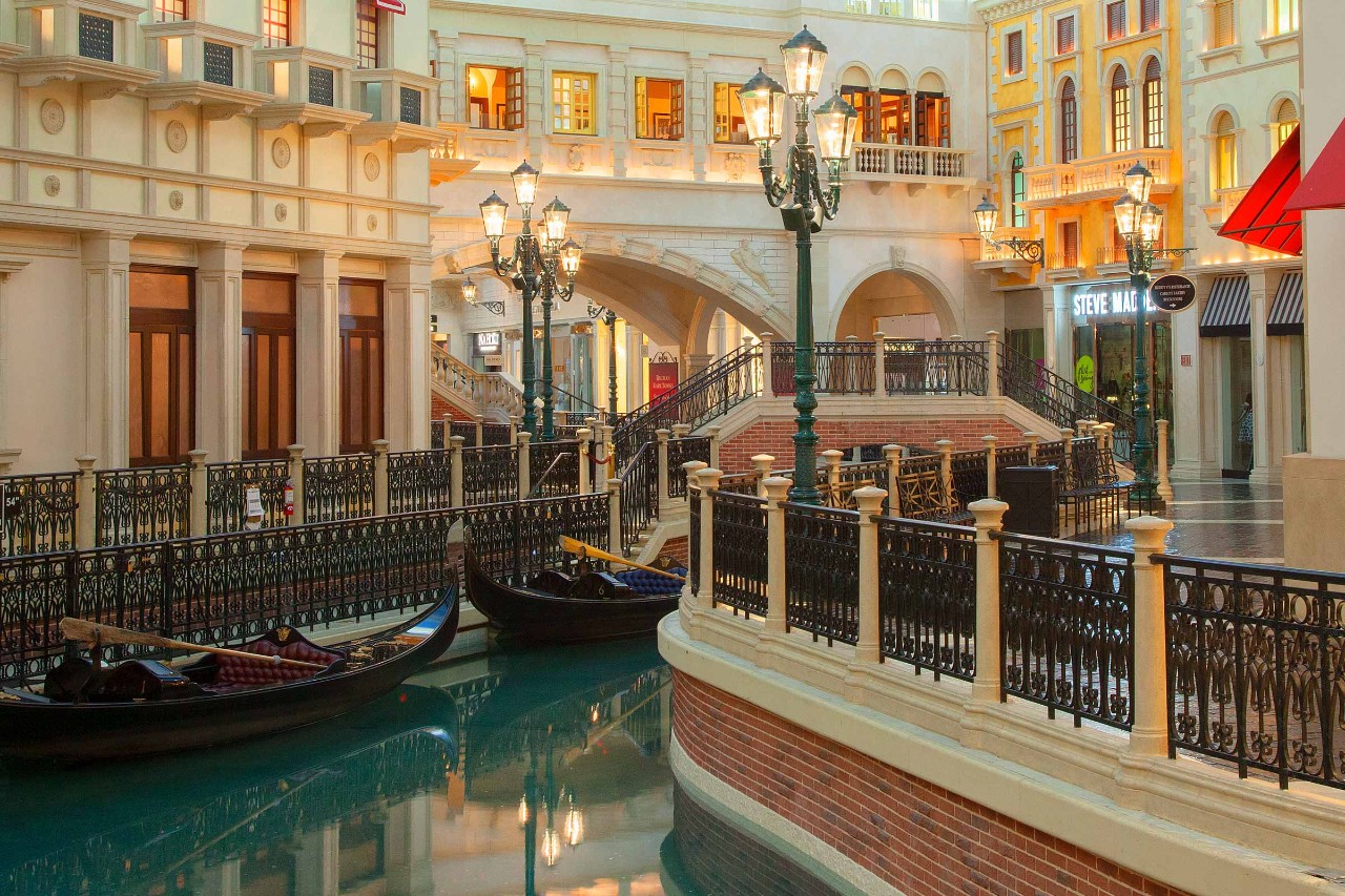 Interior of a Venetian-themed shopping mall with gondolas on a canal, ornate lampposts, and facades resembling Venice buildings.