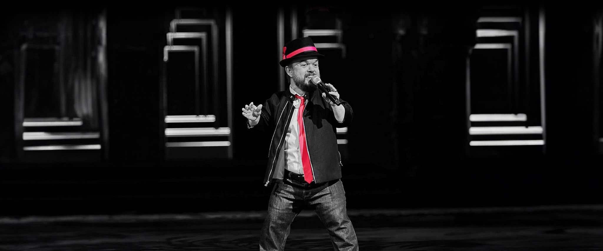 Brad Williams singing on stage in color-accented black and white, wearing a red hatband and tie.