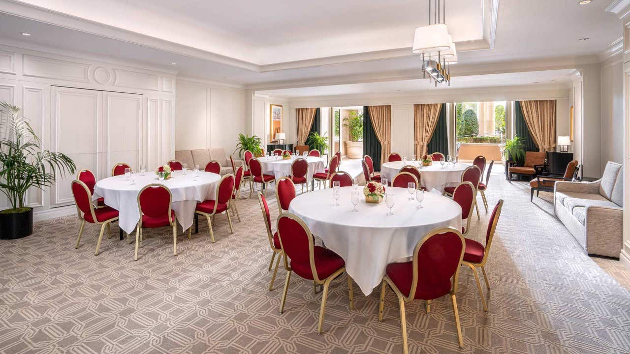 Elegant room with round tables draped in white cloth, red chairs, and large windows allowing natural light to flood in.