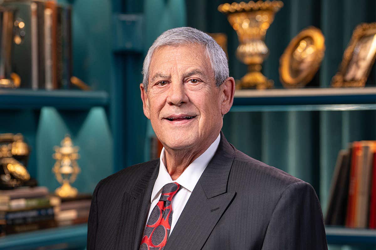 A man in a suit and tie smiles in front of a bookshelf with decorative items.