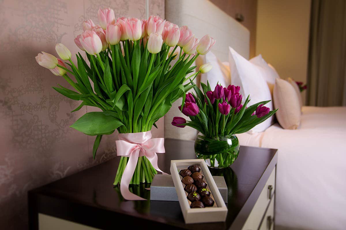 A nightstand with pink and purple tulips in vases, and a box of assorted chocolates beside a bed with white pillows.