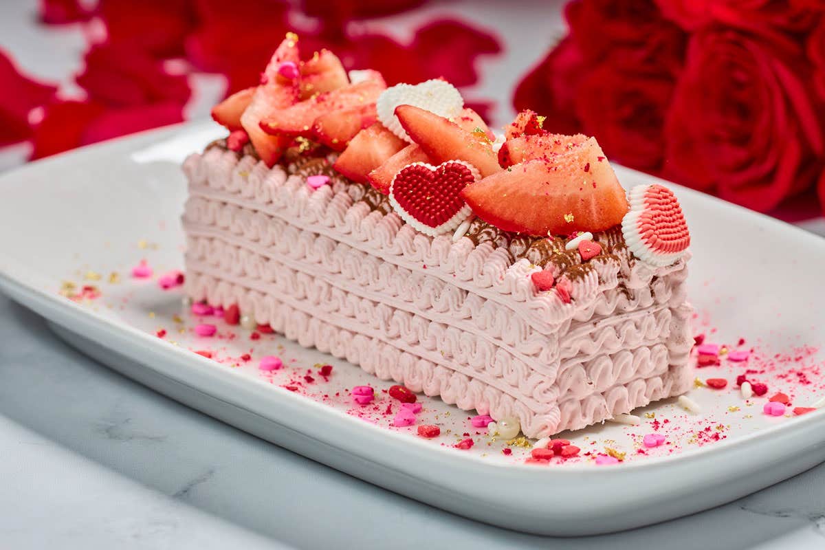 Rectangular dessert with pink whipped cream layers, topped with strawberries and heart-shaped decorations on a white platter. Roses in the background.