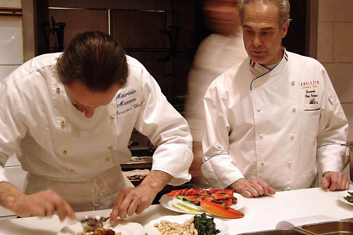 Two chefs in white uniforms plating dishes in a professional kitchen. One is focused on the food, while the other looks on.