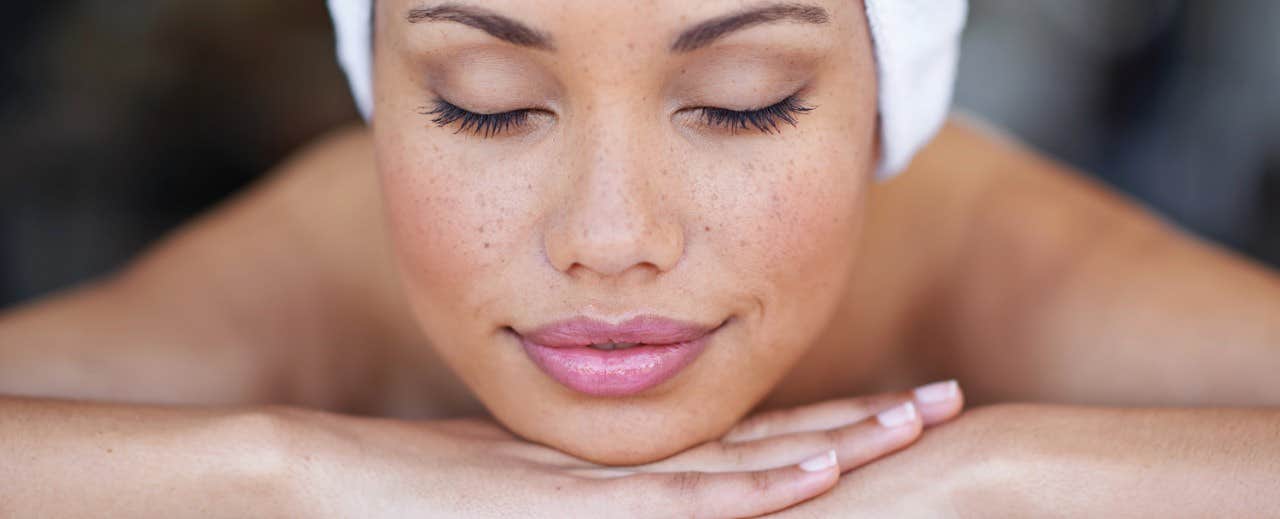 Close-up of a woman with a towel on her head, eyes closed, resting her chin on her hands, looking relaxed and serene.