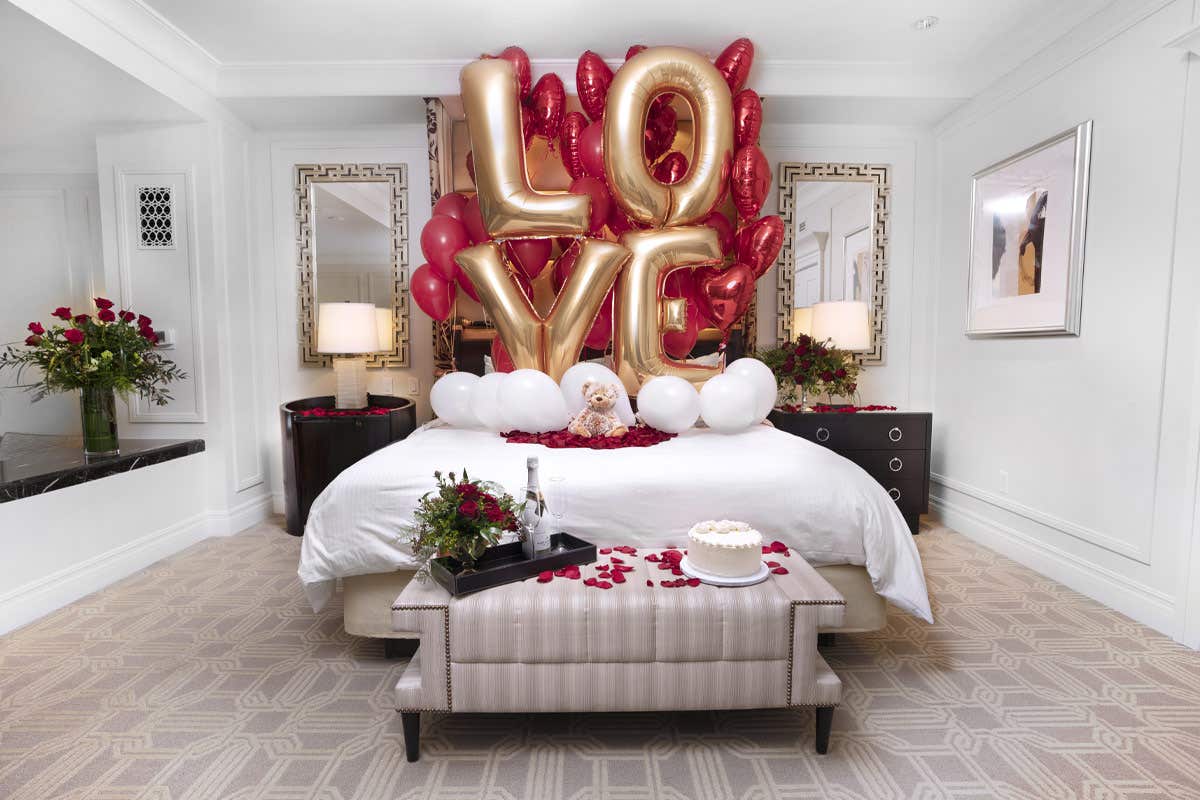 Large helium ballons spell 'LOVE' behind a large bed in a hotel suite.