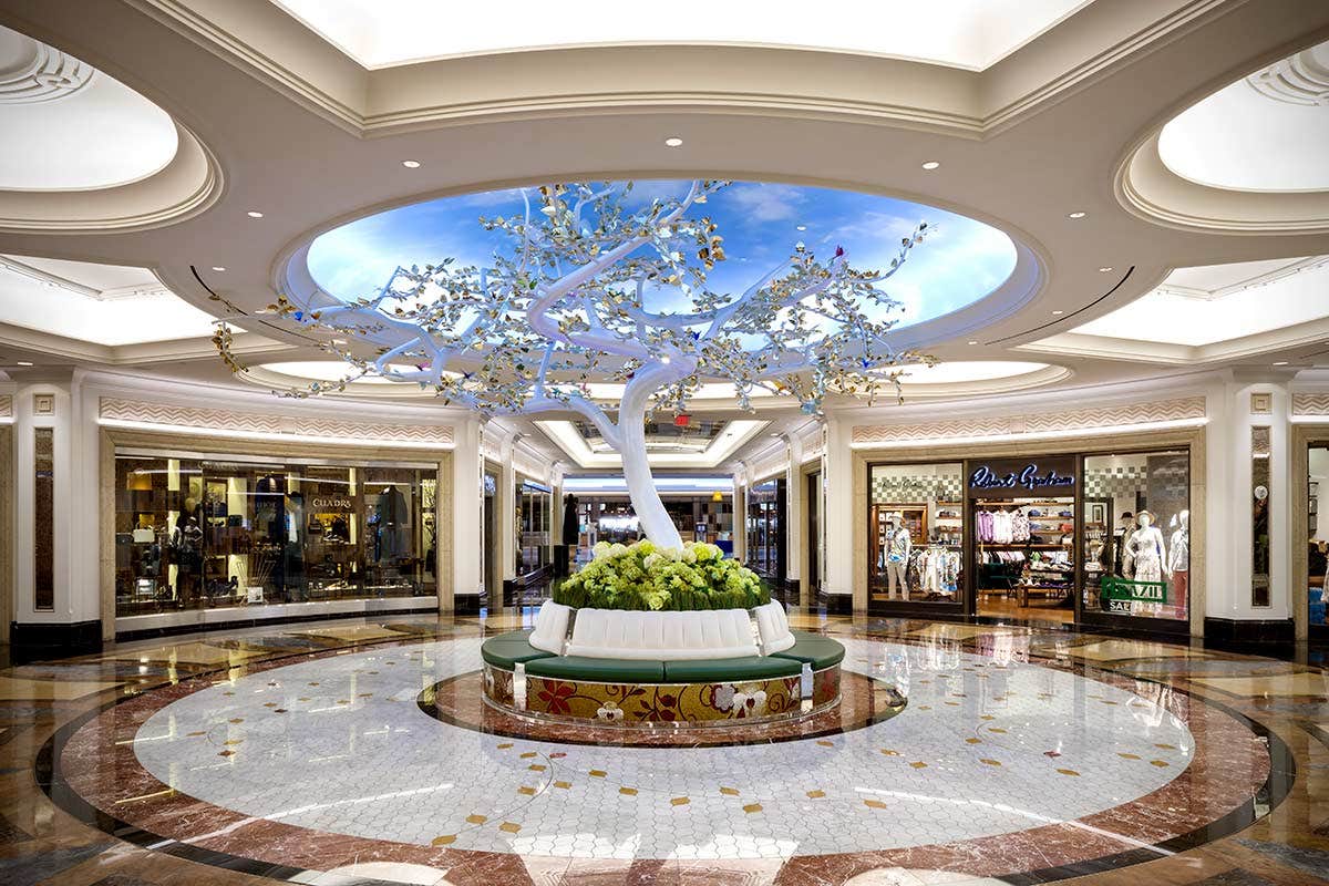 Luxurious mall interior with a white decorative tree surrounded by seating in the center and high-end stores in the background.