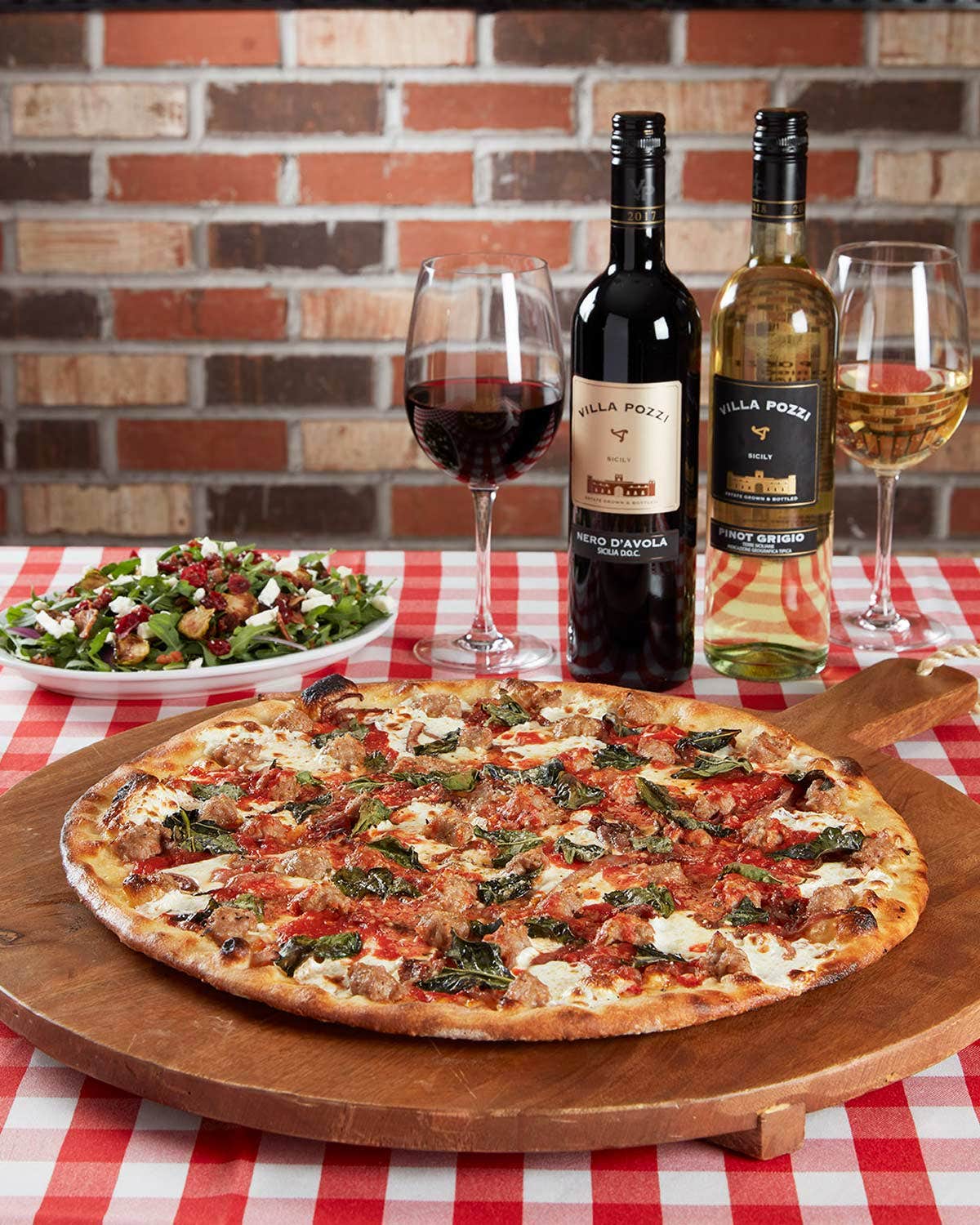 Pizza on a wooden board with a salad, two glasses of wine, and two wine bottles on a checkered tablecloth against a brick wall.