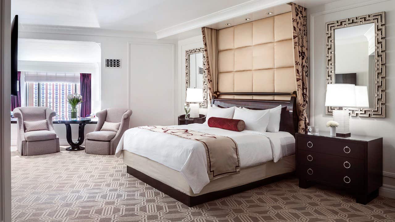 Elegantly decorated hotel room with a king-sized bed, two armchairs near a small round table, and large windows.