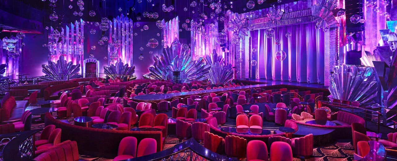 A vibrant theater with purple lighting, red chairs, and a stage adorned with crystal-like decorations and hanging spheres.