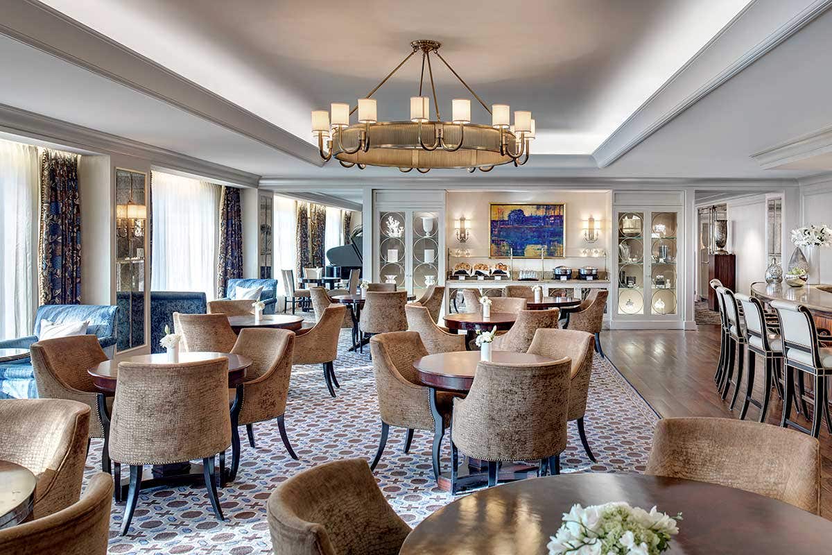 A spacious, elegant dining room with round tables, chairs, a chandelier, a bar area, and art on the walls.