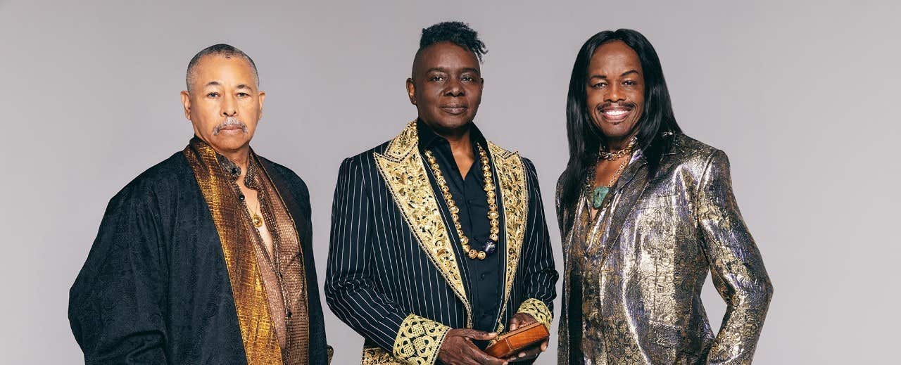 Three men in ornate jackets stand together against a neutral background, smiling and posing for the camera.