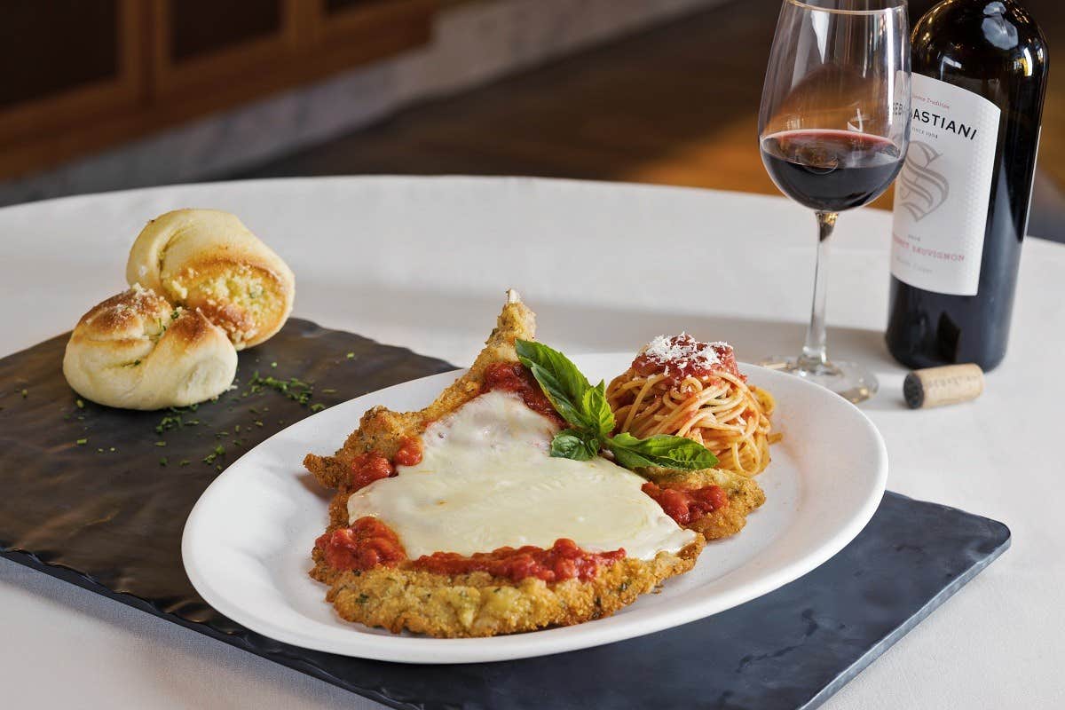 A plate of chicken Parmesan with pasta, bread rolls, a glass of red wine, and a bottle of wine on a table.