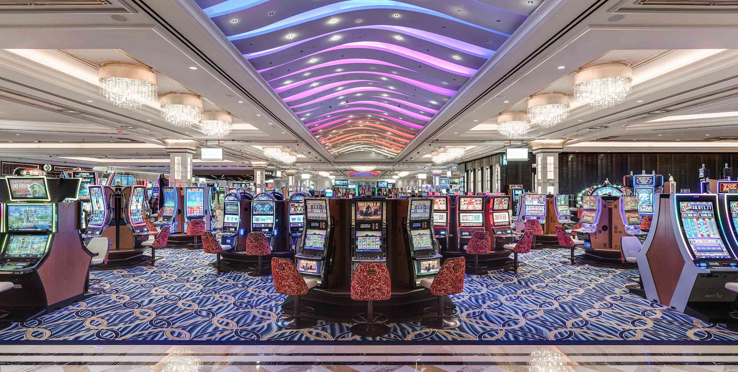 A spacious, vibrant casino featuring rows of slot machines, colorful lighting on the ceiling, and an elaborate carpet design.