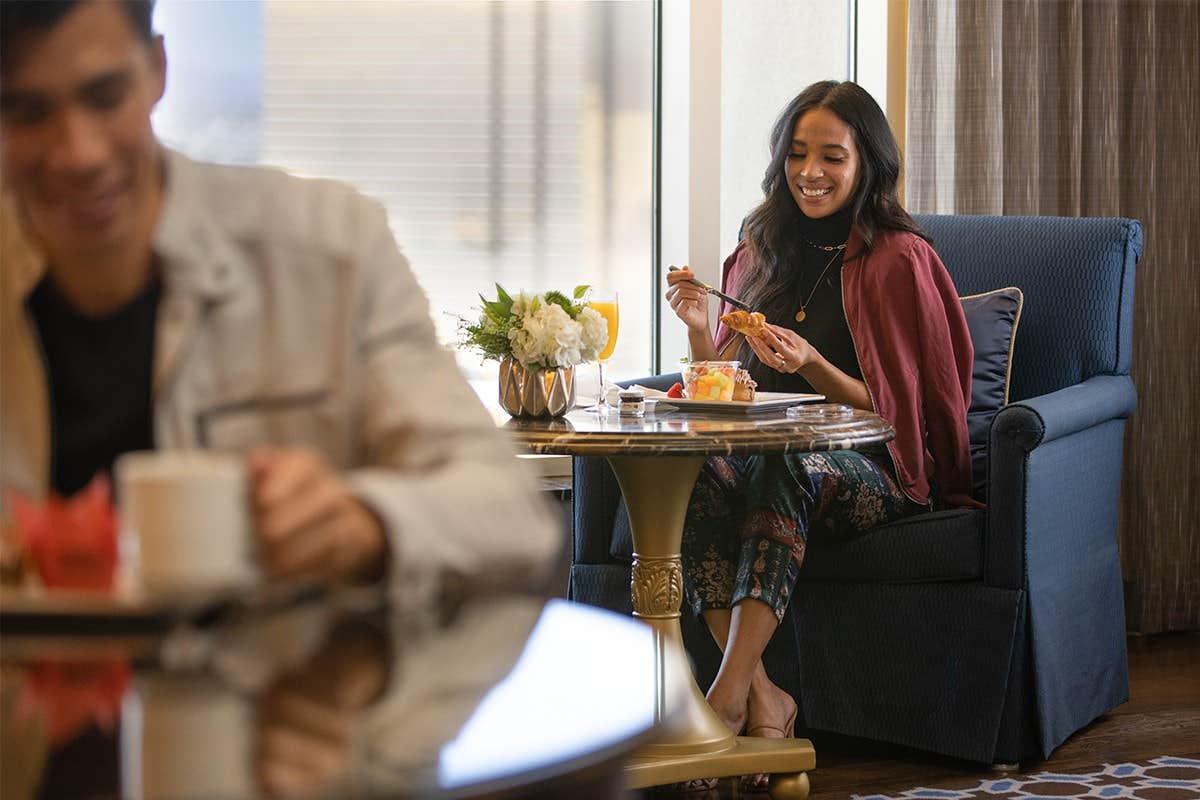 Woman happily eating at a table by a window, with a man drinking a beverage in the foreground.