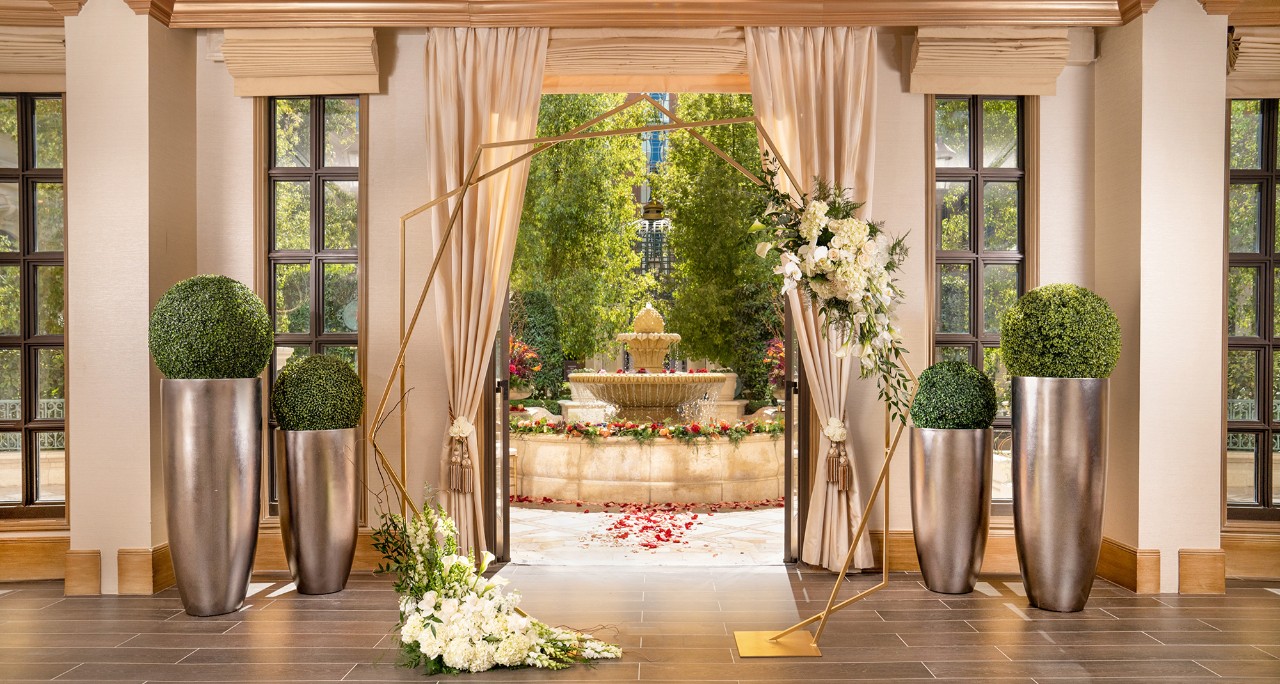 Elegant wedding arch with white flowers and greenery, facing an ornate outdoor fountain surrounded by trees and flowers.