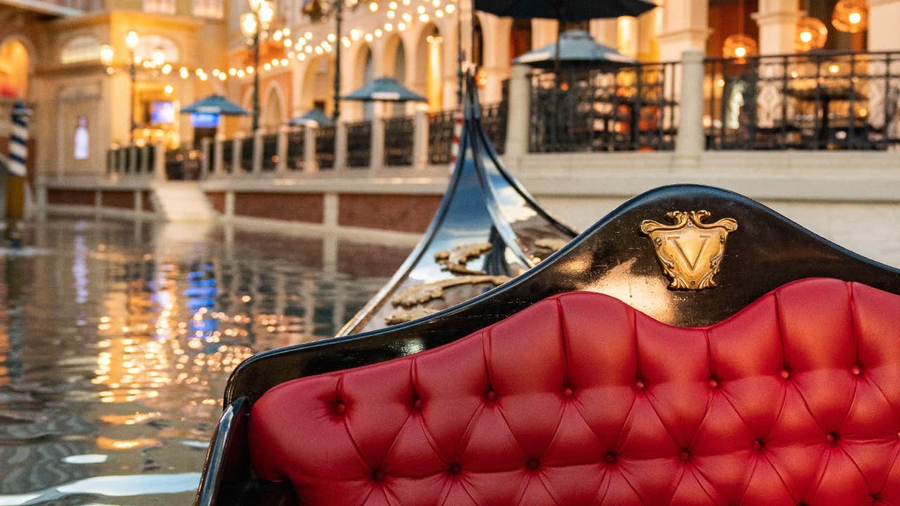 Red, tufted seat of a gondola on a canal with lit buildings and string lights in the background.