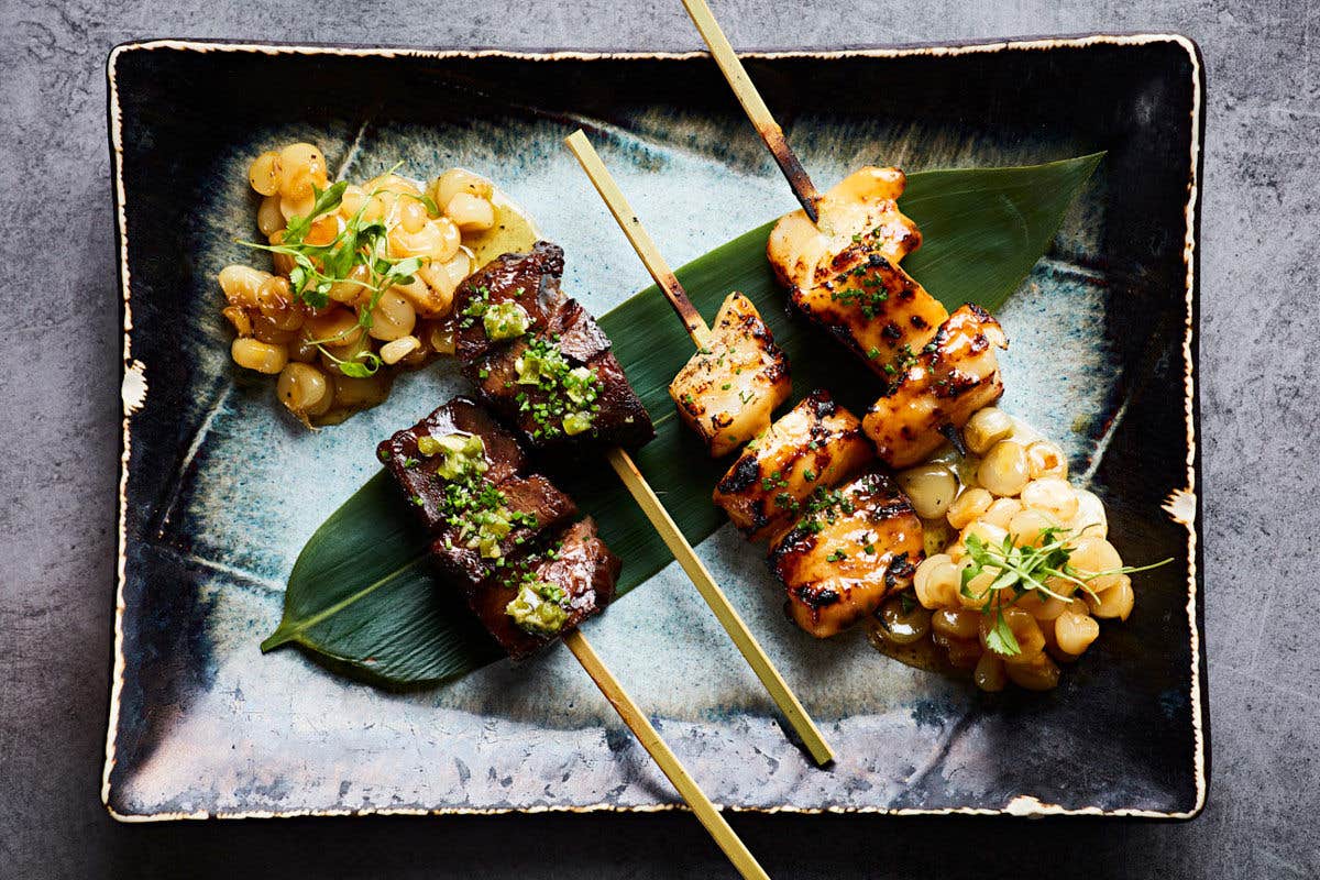 Three skewers of grilled meat and vegetables on a dark plate, garnished with fresh herbs and accompanied by cooked beans.