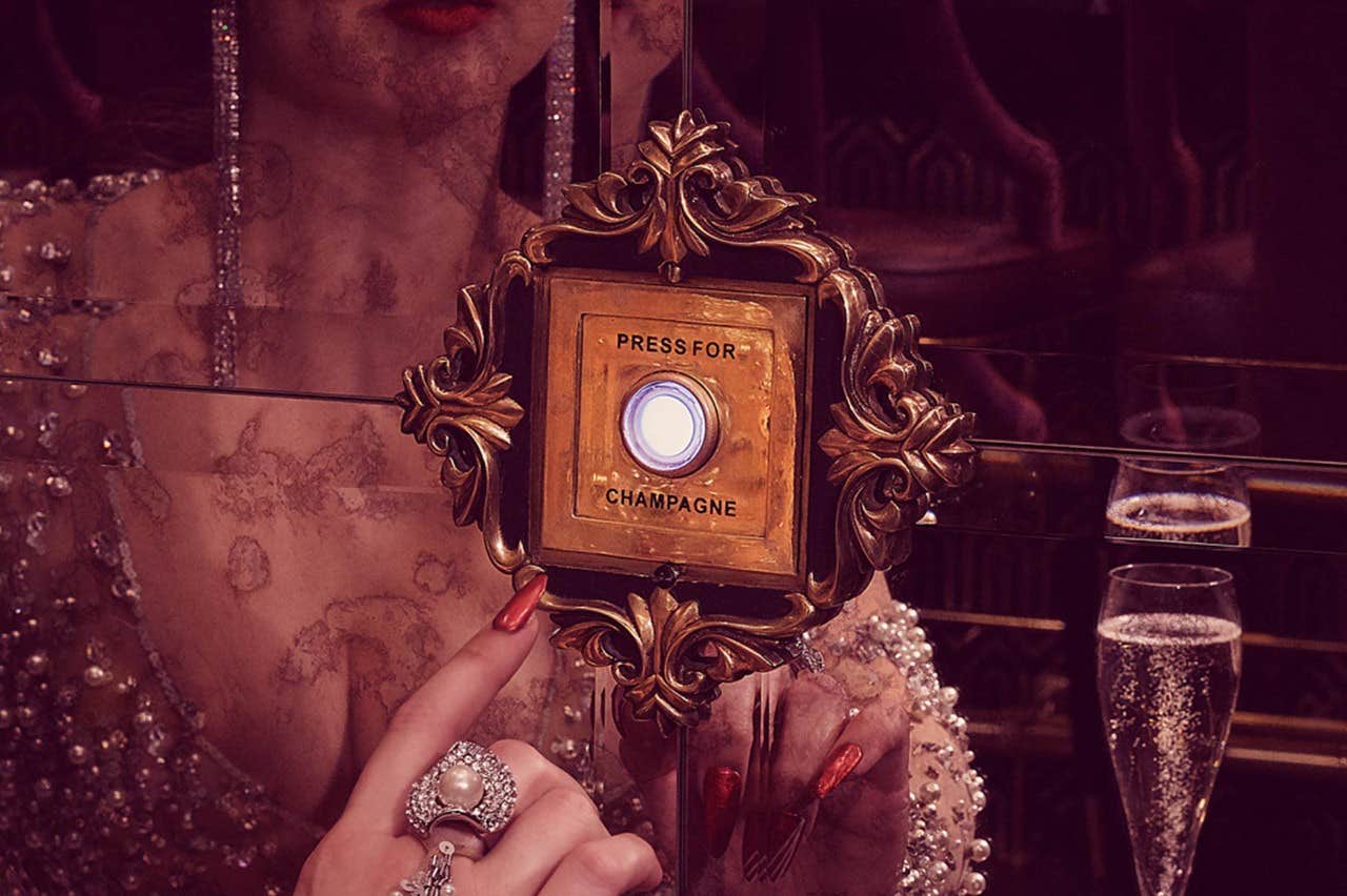 Close-up of a woman's hand pressing a button labeled "Press for Champagne" with a glass of champagne nearby.