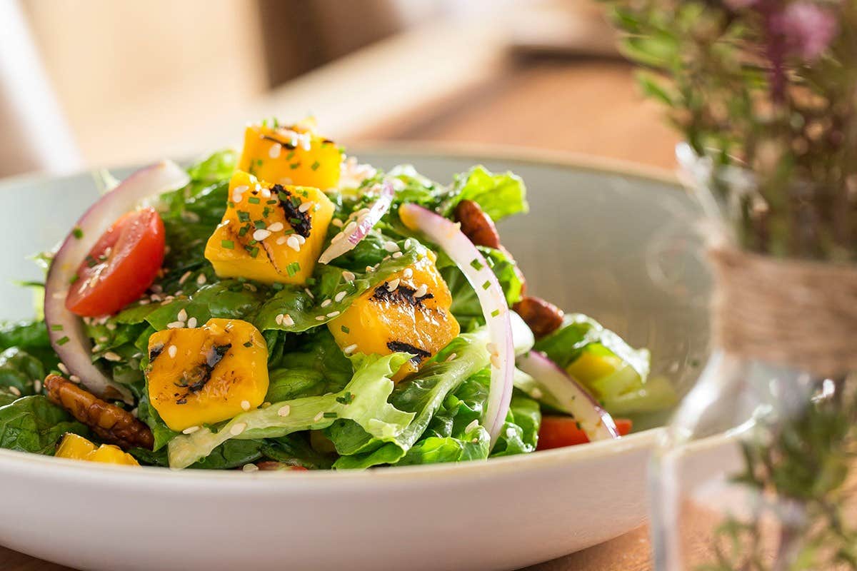 A fresh salad with lettuce, mango, cherry tomatoes, and red onions, garnished with sesame seeds in a white bowl.