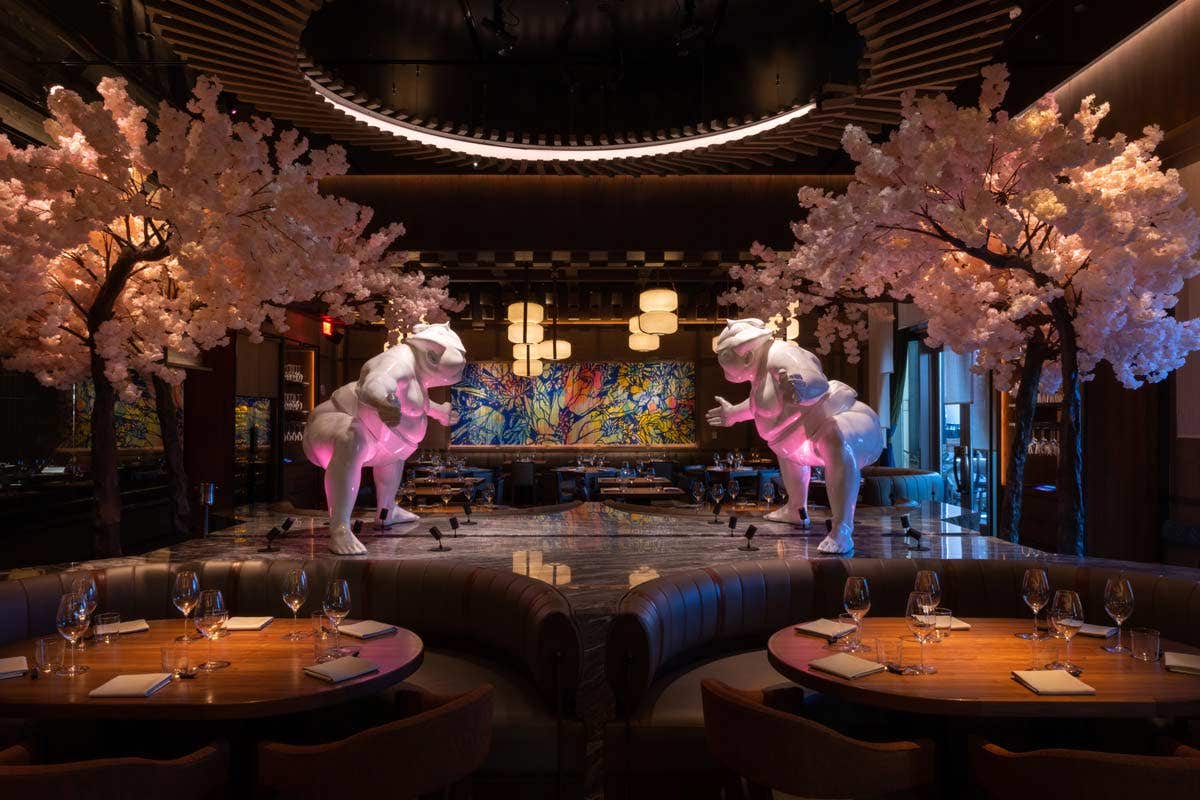 Elegant restaurant with cherry blossom trees, colorful mural, and two T-rex statues bathed in pink light.