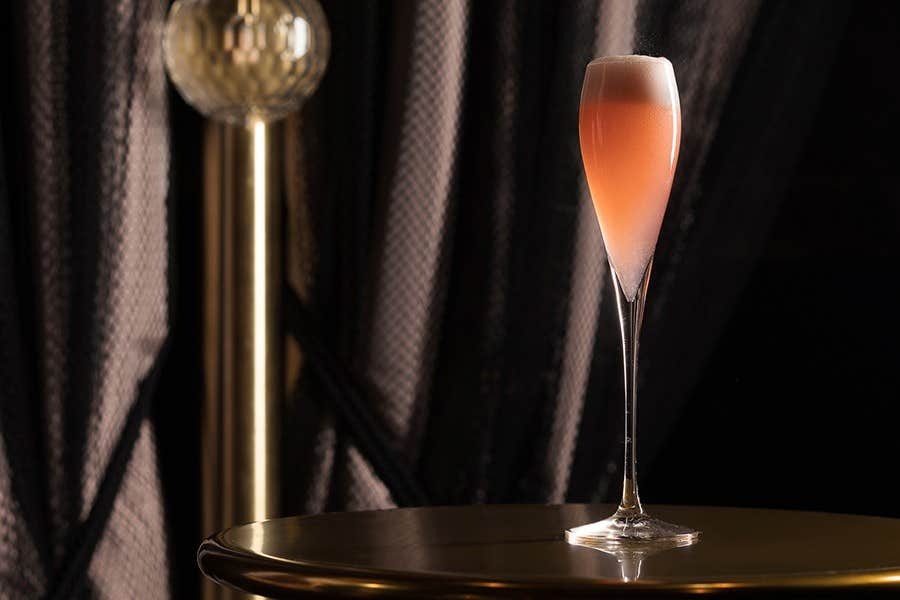 A tall champagne flute of pink cocktail on a round, shiny table with dark curtains in the background.