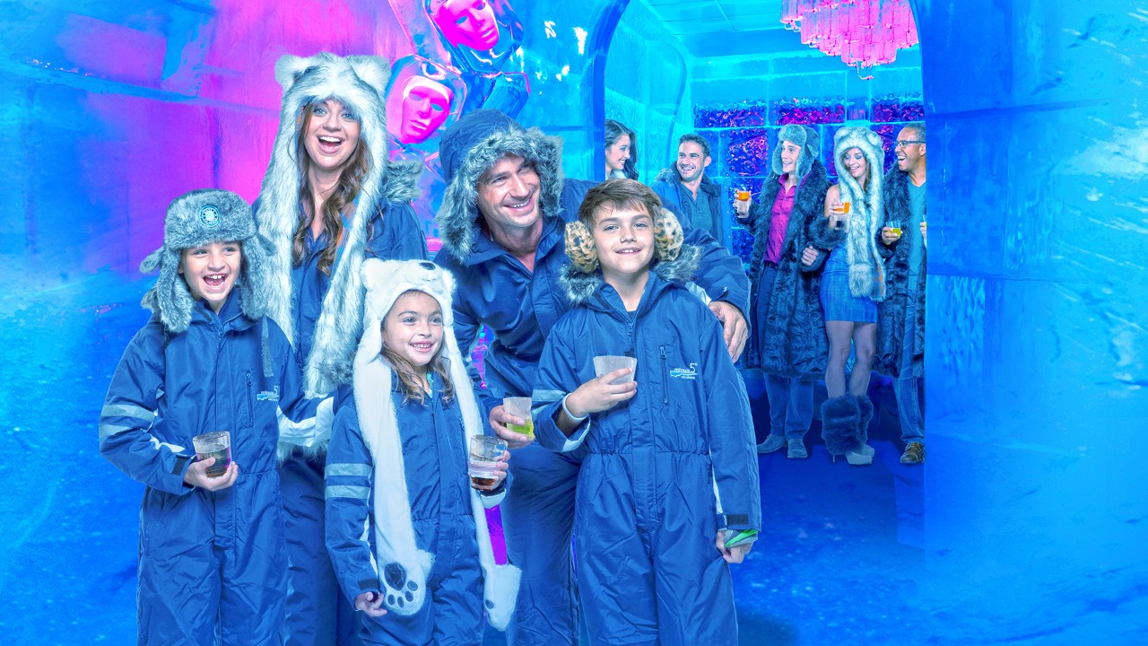 A group of people in warm blue clothing and furry hats enjoy a drink in an ice bar with blue-lit walls and ice sculptures.