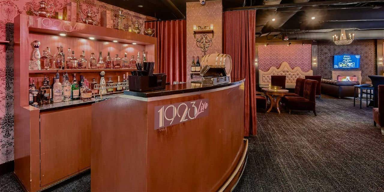 1923 Prohibition Bar entrance with a wooden bar and decorated shelves, set against floral wallpaper and plush seating.