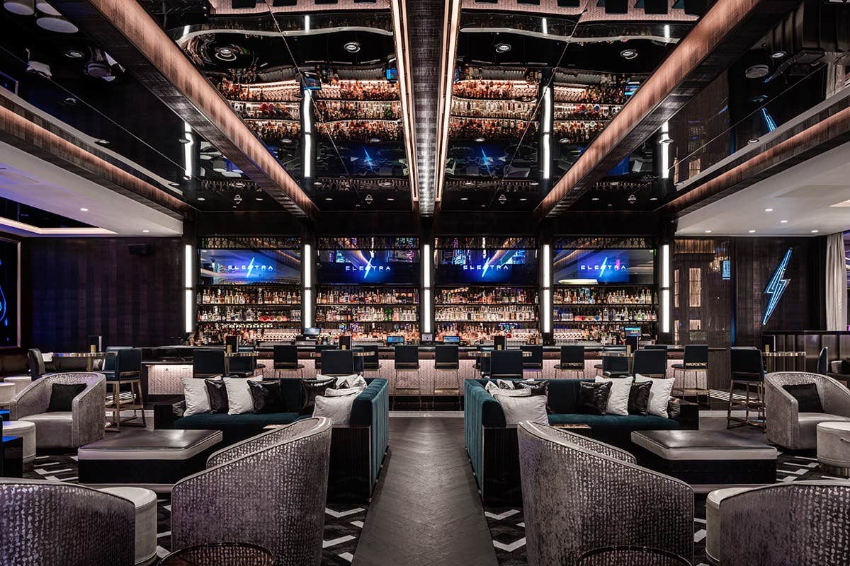 Modern upscale lounge with plush seating, a long central bar, mirrored ceiling, and multiple shelves of bottles behind the bar.