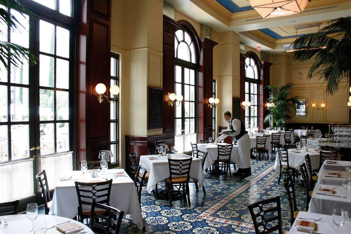 A waiter sets tables in an elegant restaurant with large windows, tiled floor, and white tablecloths.