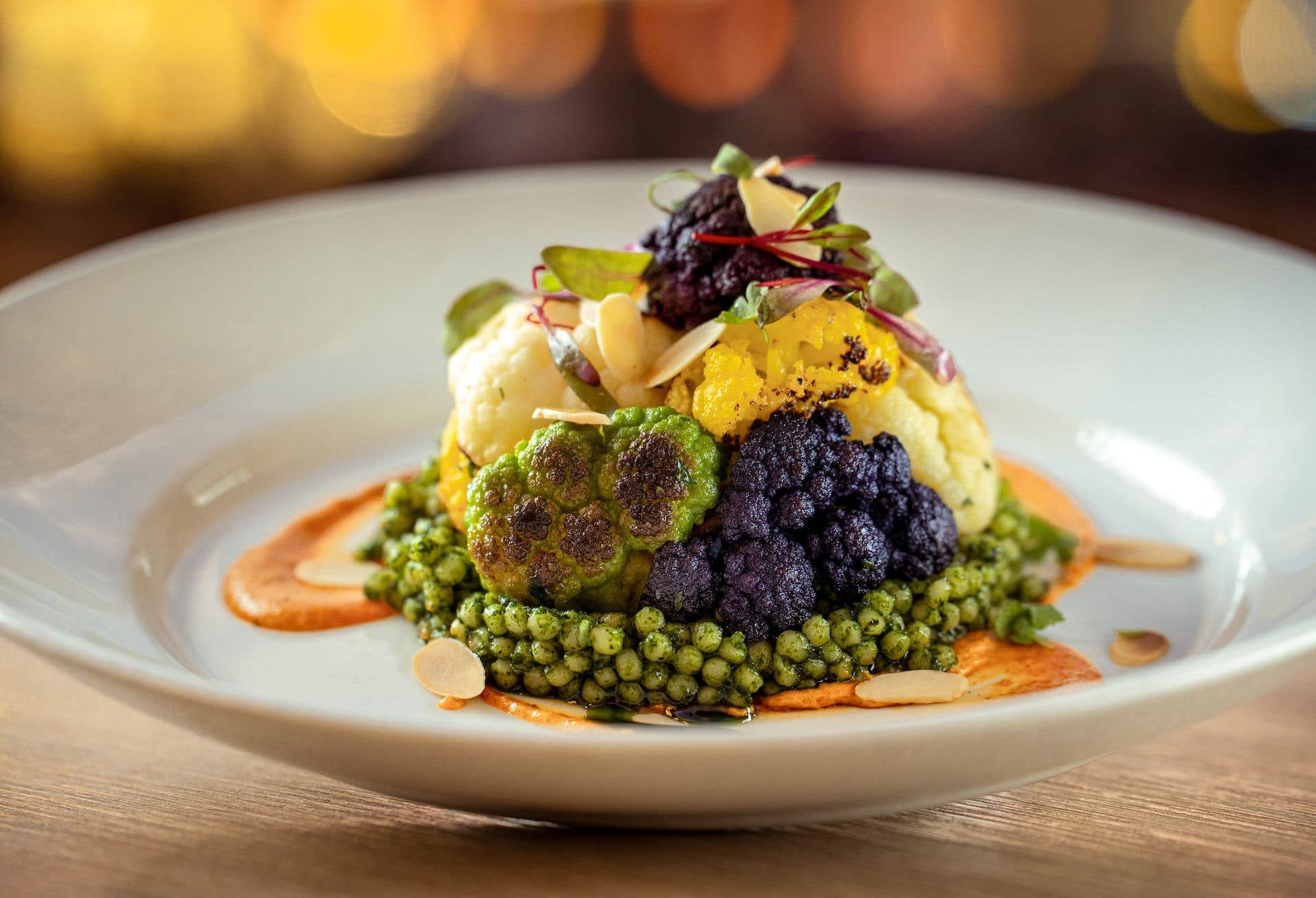 A dish of roasted multi-colored cauliflower on top a romesco sauce, garnished with micro greens and slivered toasted almonds.