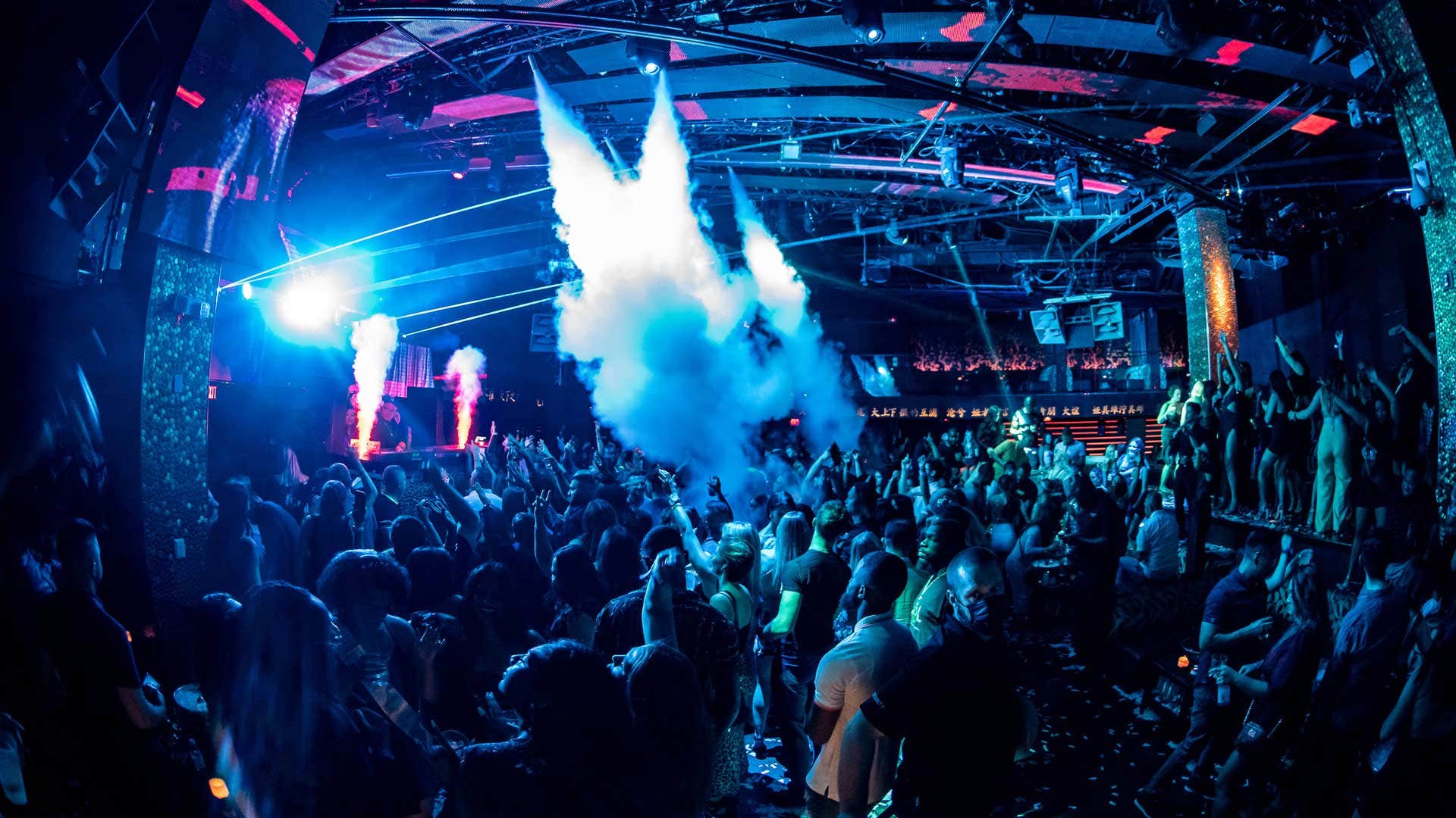 A crowded nightclub with colorful lights and beams illuminating people dancing.