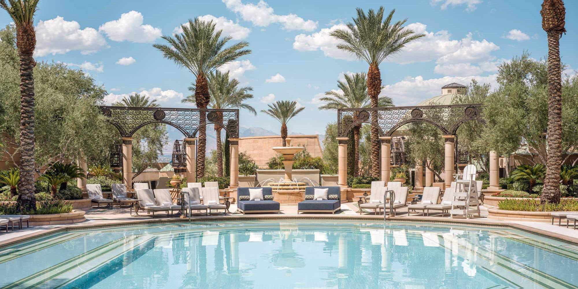 A luxurious swimming pool with lounge chairs, palm trees, and a lavish resort building in the background on a sunny day.