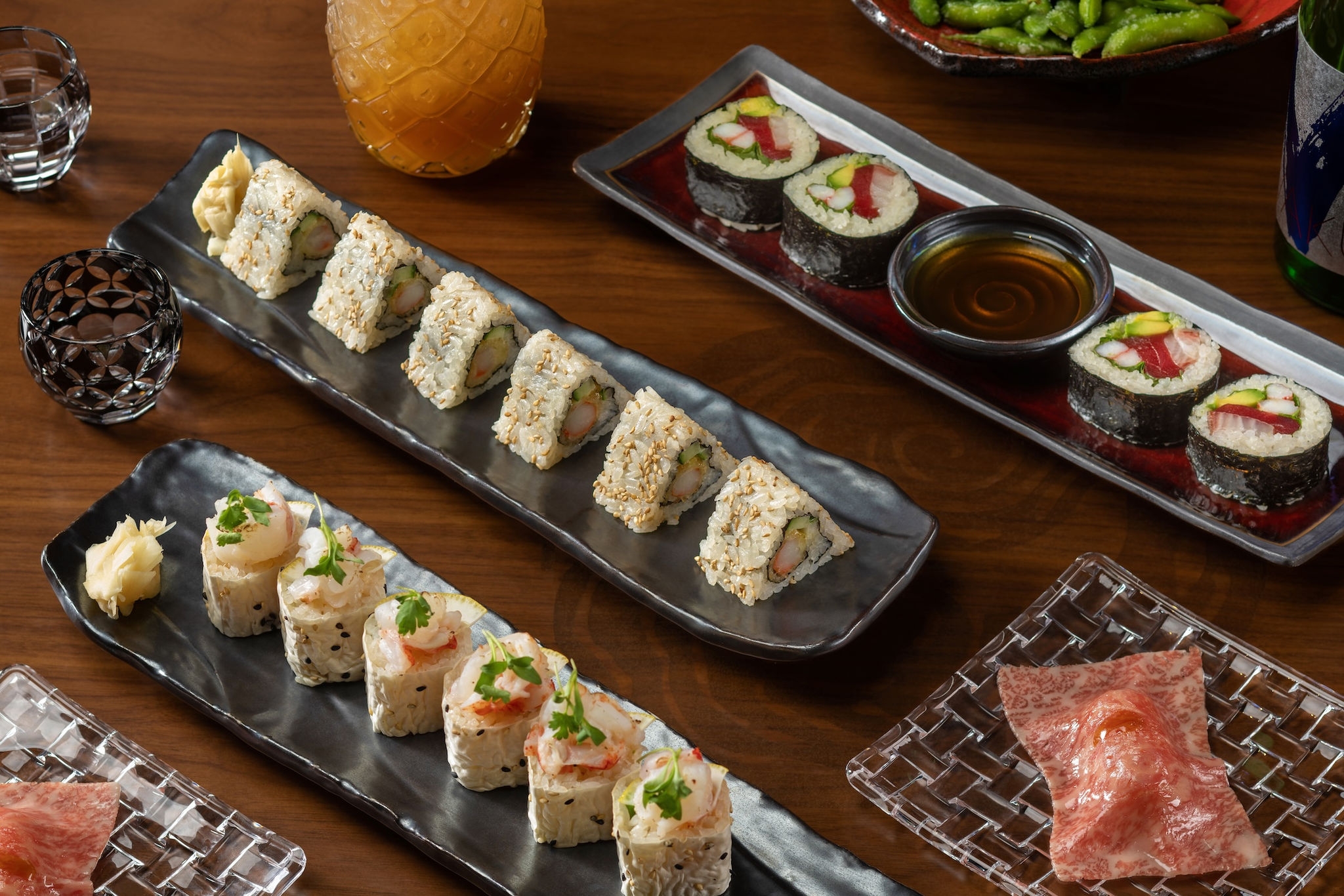 A wooden table with assorted sushi rolls, a drink, soy sauce, edamame, and garnish on various plates.