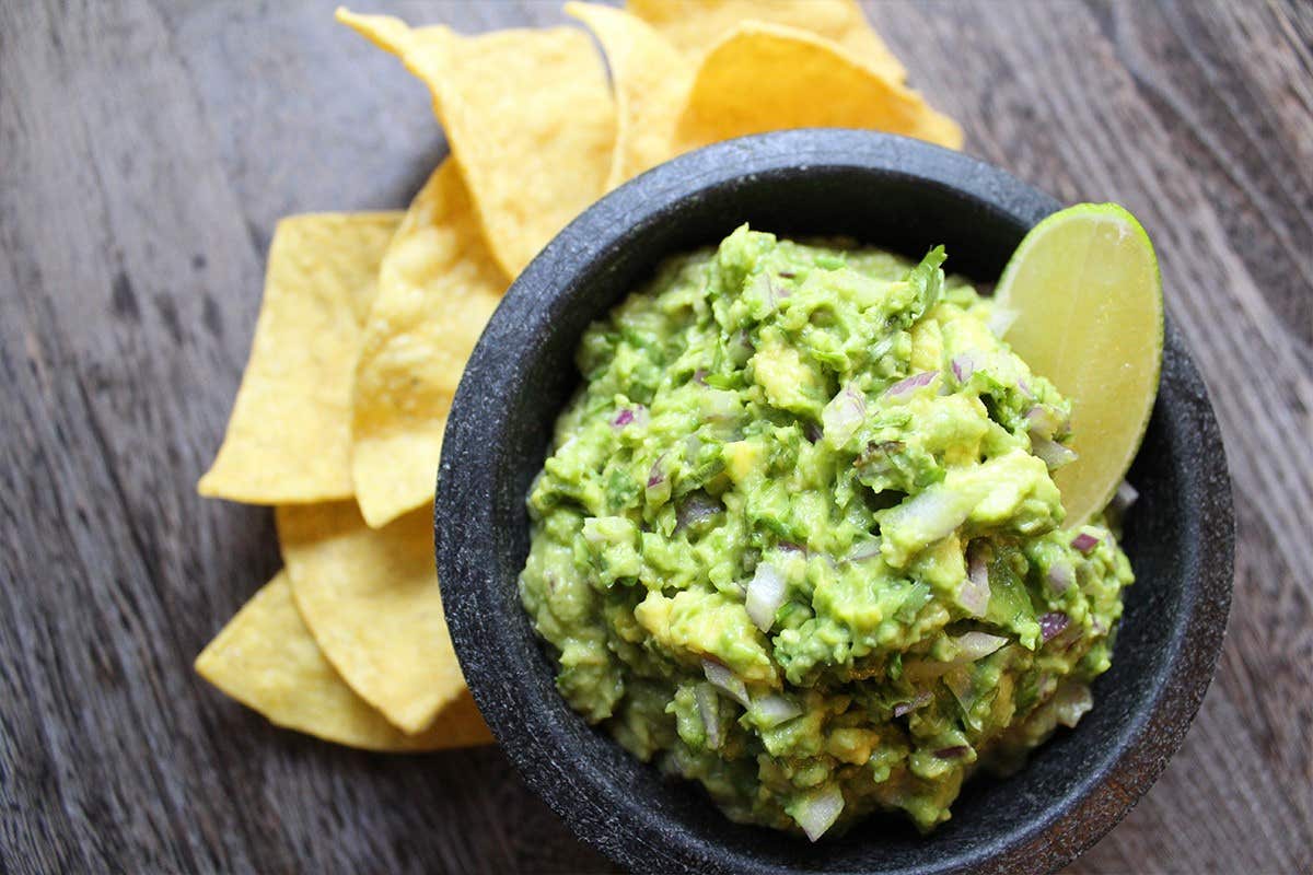 Bowl of guacamole garnished with a lime wedge, surrounded by tortilla chips on a wooden surface.