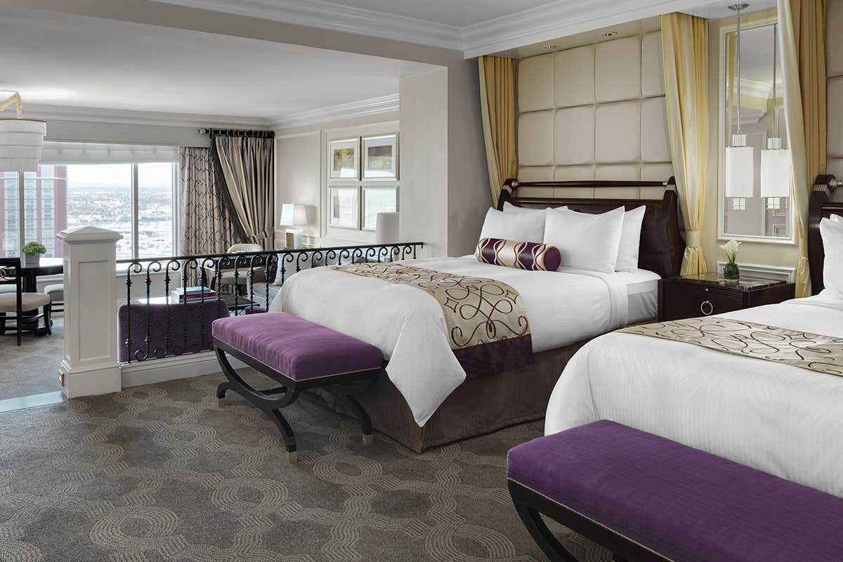 Luxurious hotel room with two large beds, purple accents, sitting area, and a city view. Beige and purple color scheme.