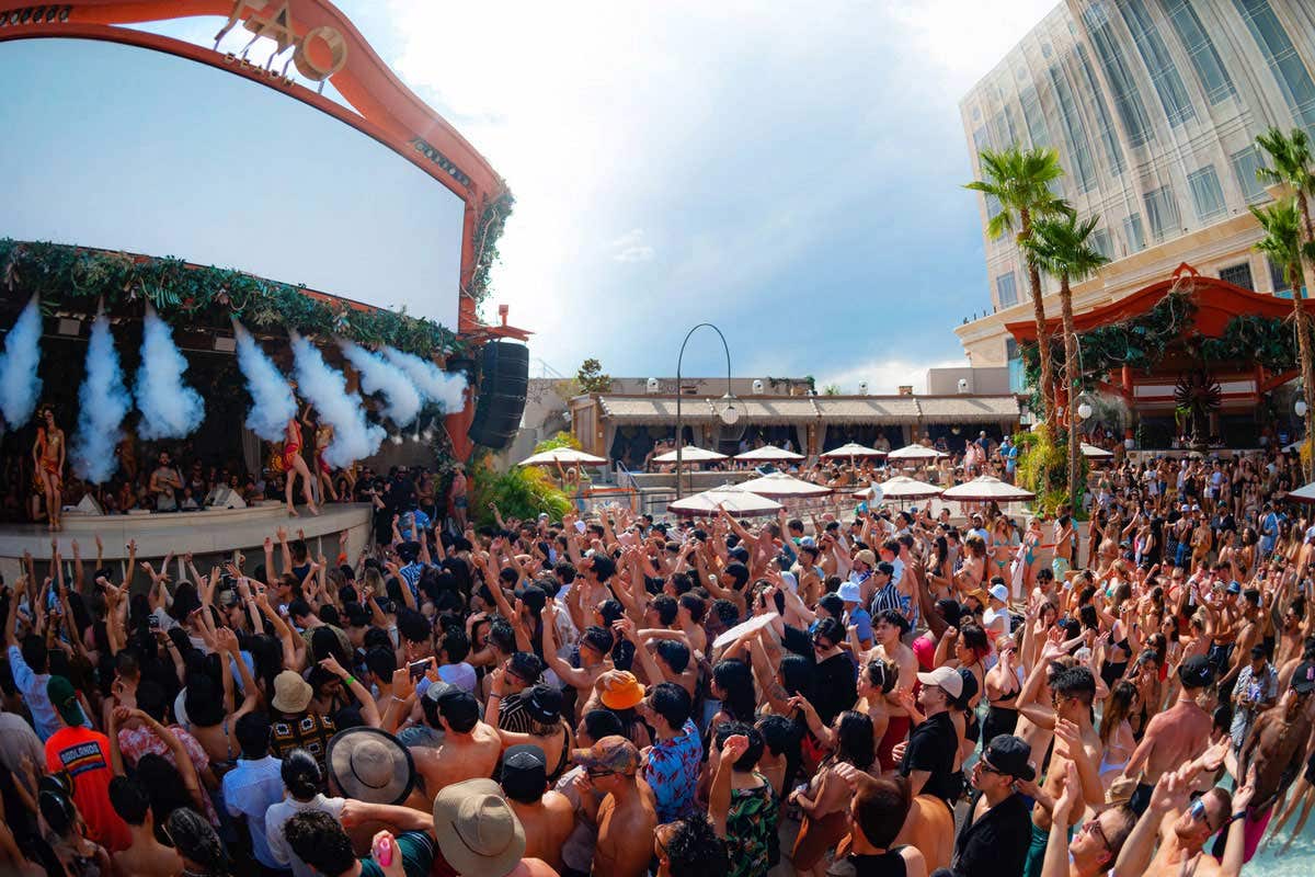 A large crowd at TAO Beach, with palm trees to the right, and on the left, the DJ booth with multiple streams of fog machines on