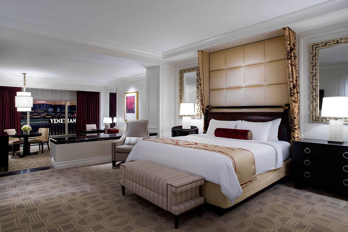 Luxurious hotel room with a large bed, ornate mirrors, a lounge chair, and a view of the "VENETIAN" sign through the window.