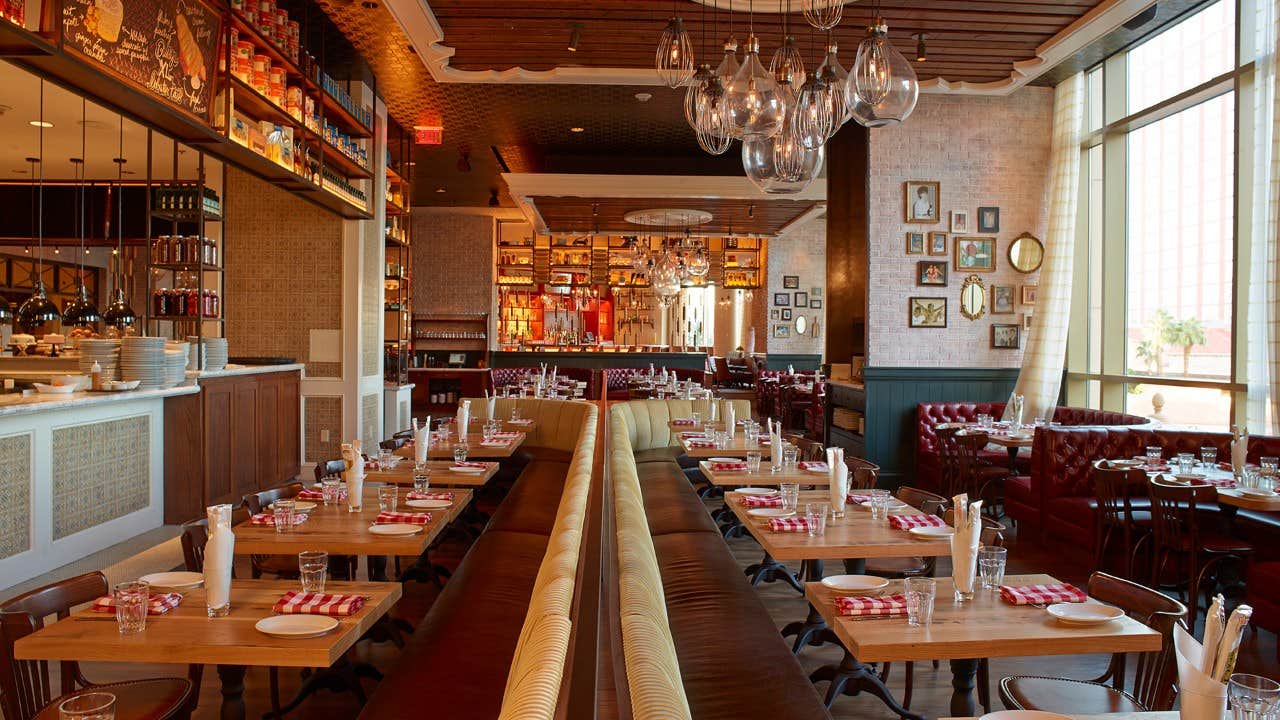 A warmly lit restaurant with wooden tables, red leather benches, hanging lights, and a bar in the background.