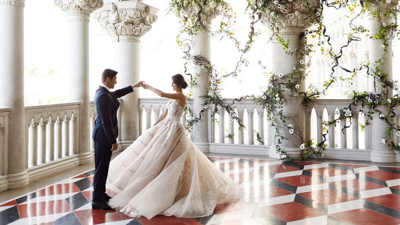 A couple elegantly dances on a checkered floor in a grand hall adorned with columns and climbing greenery.