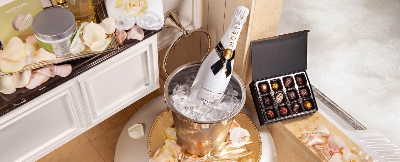 Champagne in an ice bucket, chocolates in a box, and rose petals on a bath tray next to a bubble-filled bathtub.